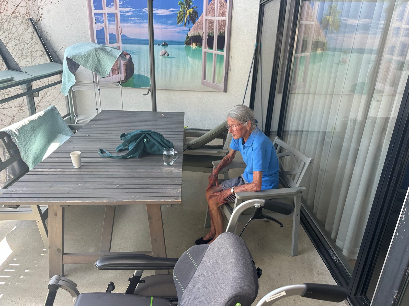 Trudy, zittend op een besloten balkon in de zorginstelling, met op de achtergrond een muurposter van een tropisch strand.